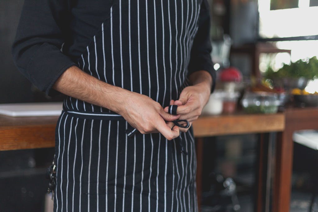 Chef tying a black and white striped apron in an indoor kitchen with fresh ingredients nearby.