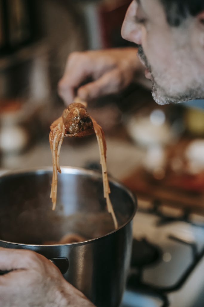 Crop middle aged ethnic male cook blowing on delicious saucy pasta with meatballs prepared in metal saucepan in kitchen